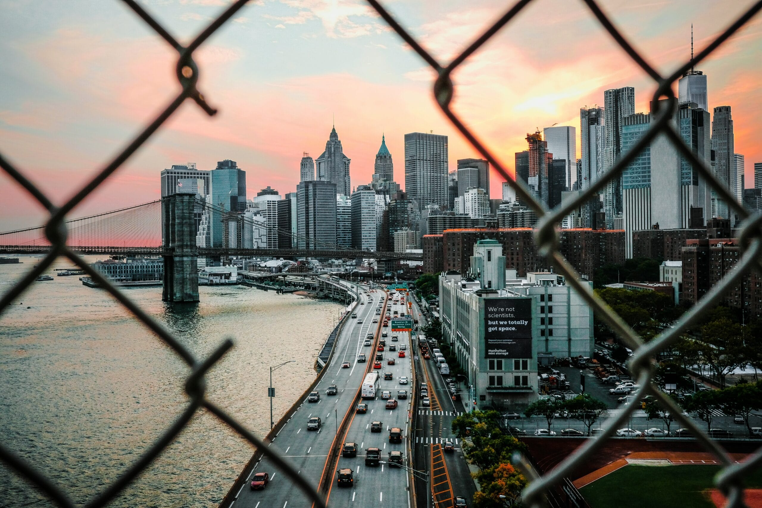 A city skyline with tall buildings at sunset, viewed through a chain-link fence, with a river, bridge, and busy road with cars in the foreground.