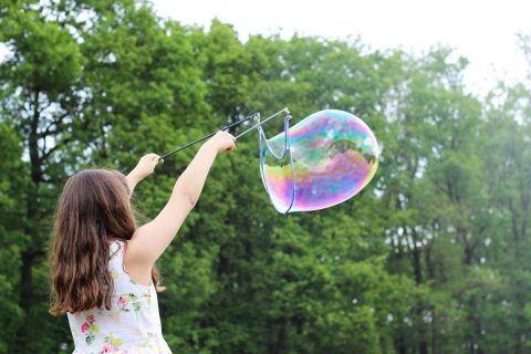 A girl in a floral dress creates a large soap bubble outdoors using a bubble wand, with green trees in the background.