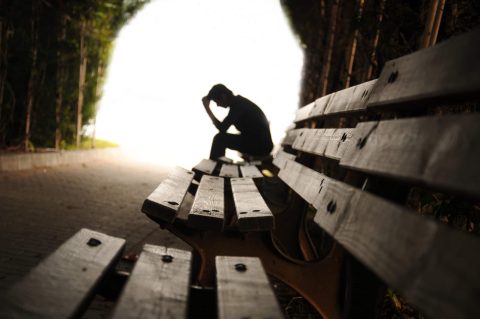 A person sits on a bench with head in hand, silhouetted against a bright background at the end of a tree-lined path.