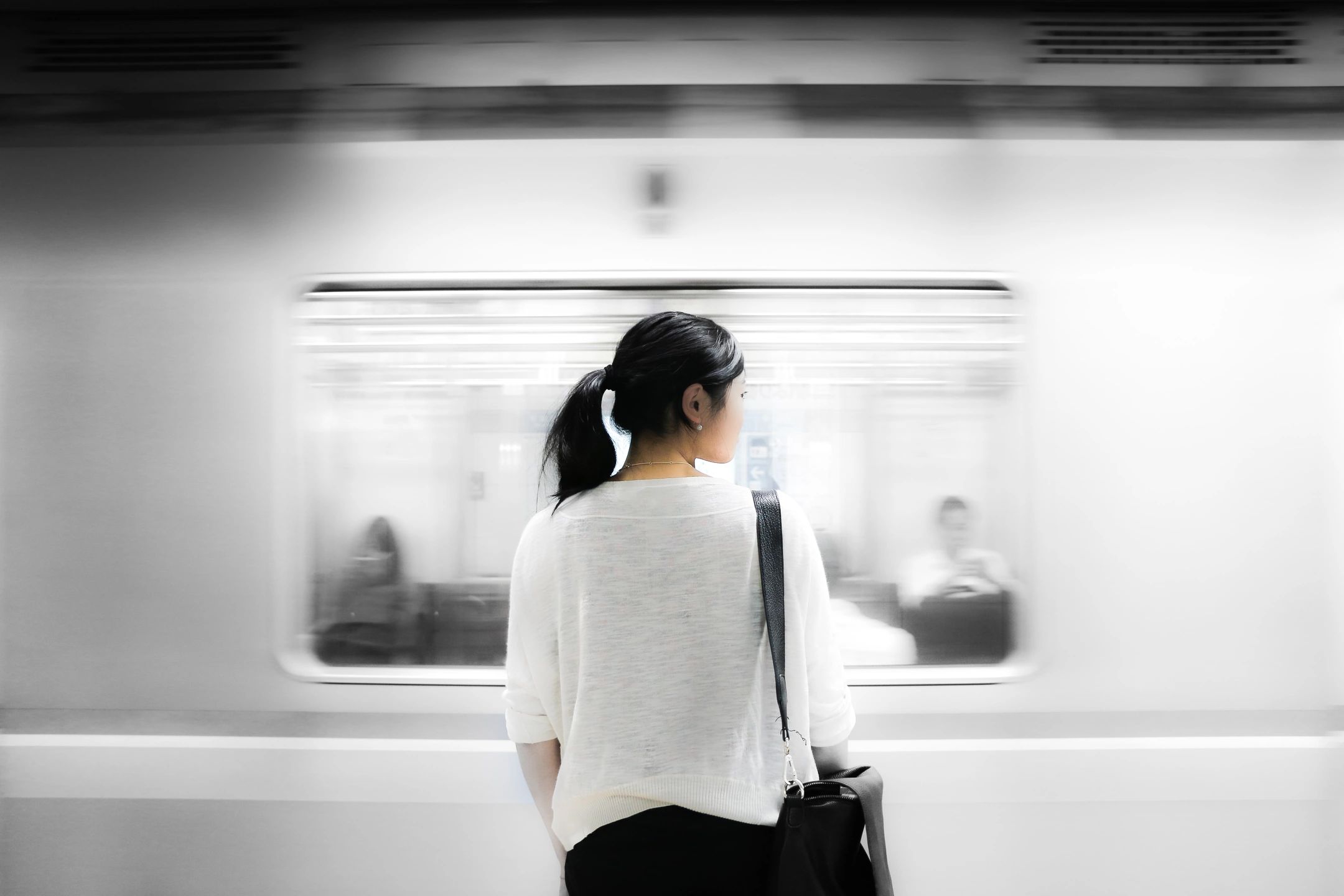 A person with a ponytail stands on a subway platform, facing a passing train, with blurred motion and seated passengers visible through the window.