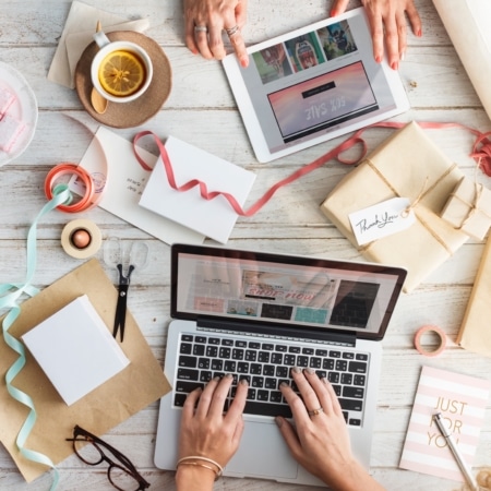 Two people work on gift wrapping at a white wooden table with a laptop, tablet, ribbon, gift boxes, envelopes, and a cup of tea.