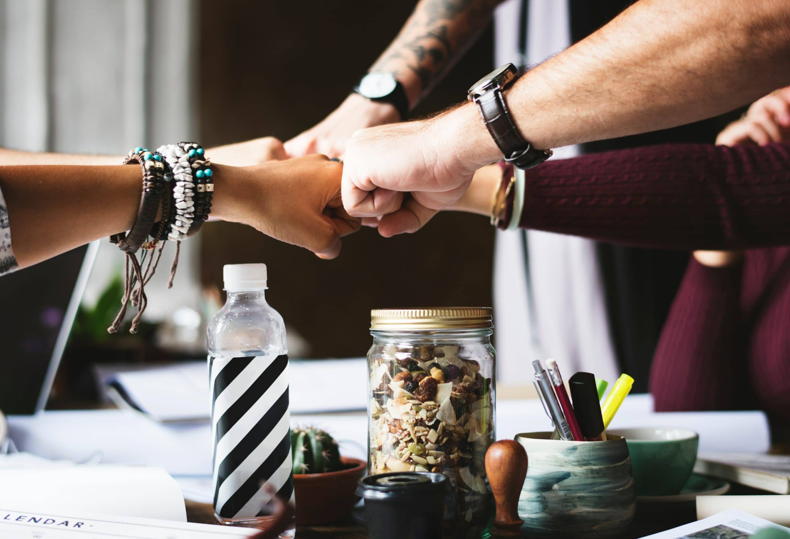 Four people perform a group fist bump over a desk with a water bottle, jar of mixed nuts, papers, and office supplies.