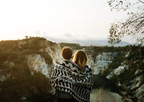Two people wrapped in a patterned blanket stand together, facing a scenic view of rocky cliffs and a river or lake, with trees and a hazy sky in the background.