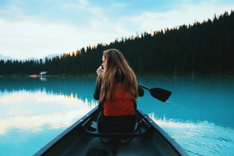 A person with long hair in an orange life jacket sits in a canoe on a calm blue lake, paddling toward a forested shoreline at sunset.