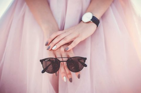 Close-up of a person’s hands with painted nails resting on a pink skirt, holding black sunglasses, and wearing a wristwatch with a black strap.