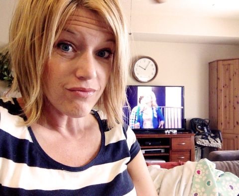 A woman with blonde hair in a striped shirt sits indoors; a clock and TV showing two people are visible in the background.