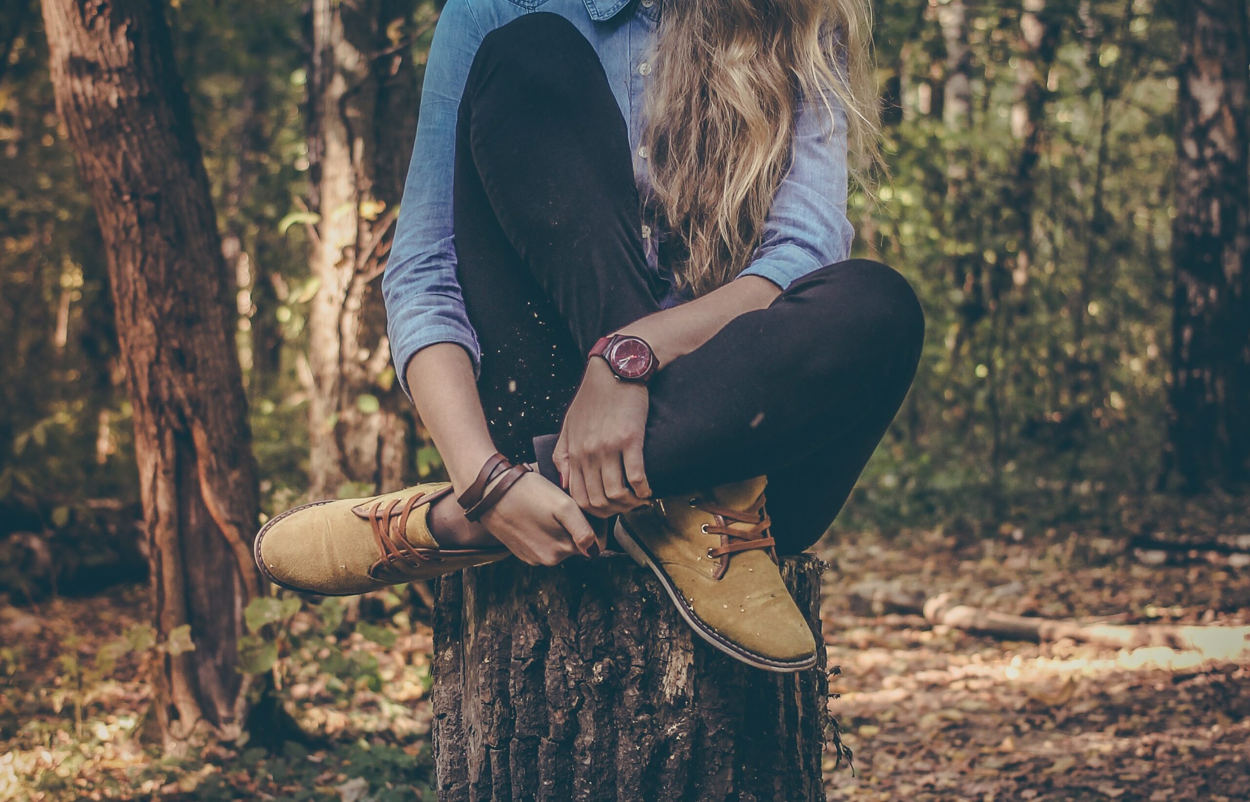 A person with long hair sits cross-legged on a tree stump in a forest, wearing a blue shirt, black pants, yellow shoes, and a watch.