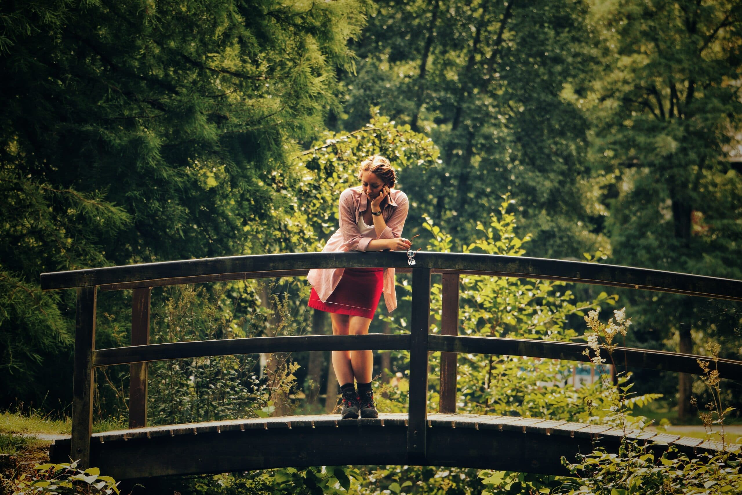 A person in a red skirt and light jacket stands leaning on the railing of a small wooden bridge in a green park.