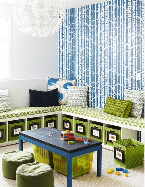 Children’s playroom with a blue and white birch tree wallpaper, L-shaped cushioned bench seating, storage bins, a blue table with toys, and green poufs.