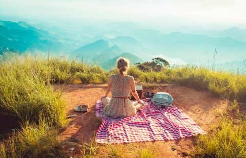 A person sits on a red and white checkered blanket atop a grassy hill, facing distant mountains under a hazy sky. Shoes and a backpack are nearby.