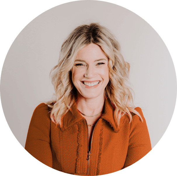 Smiling woman with wavy blonde hair wearing an orange jacket, posing against a plain light background.