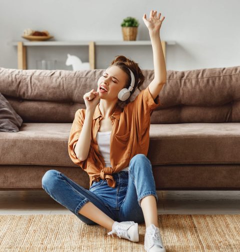 Young woman sitting on the floor in front of a sofa, wearing headphones, with eyes closed and one arm raised, appearing to sing or enjoy music.