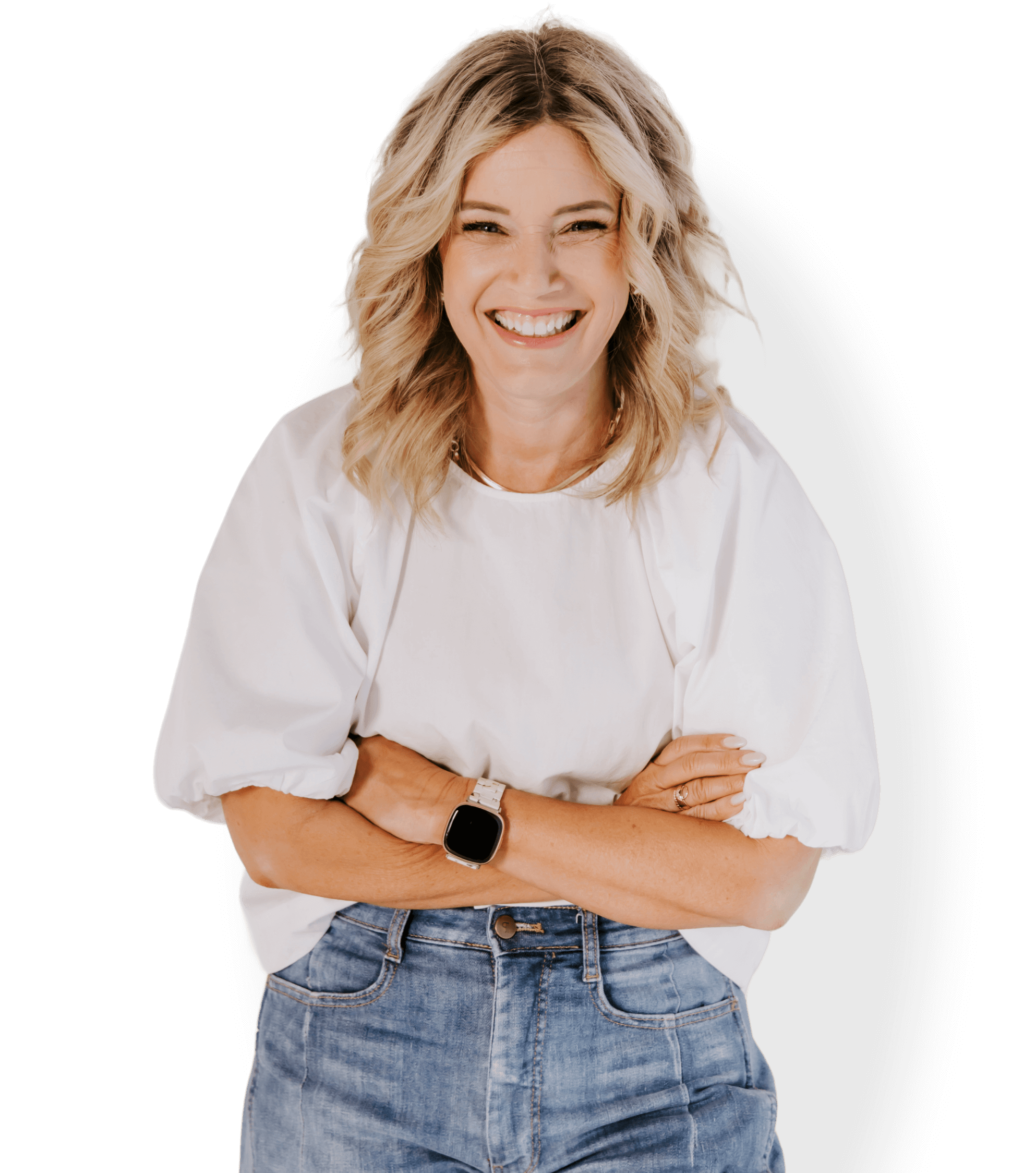 A woman with blonde wavy hair wearing a white blouse, blue jeans, and a smartwatch, standing with arms crossed and smiling.