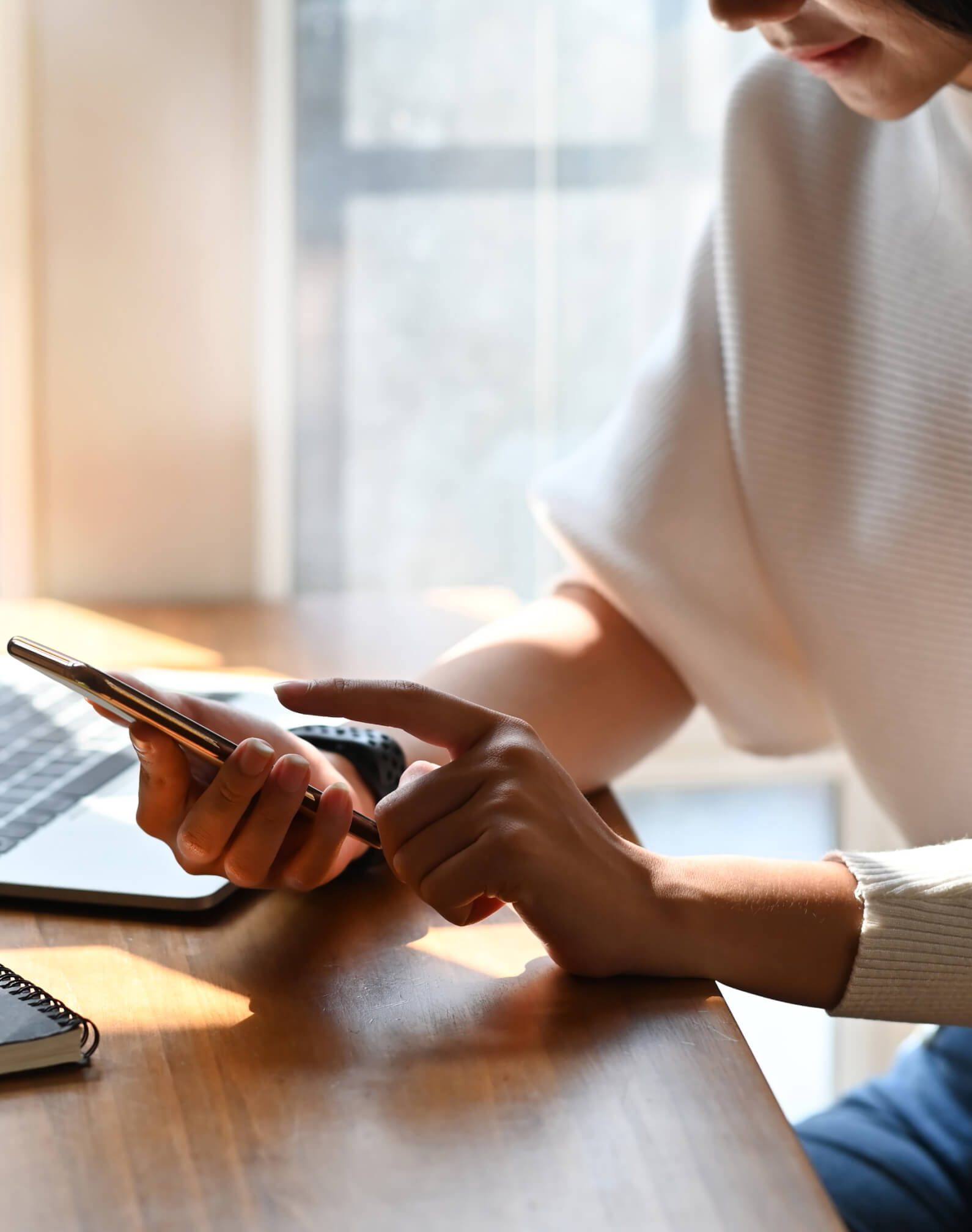 Person sitting at a desk using a smartphone, with a laptop, notebook, and pen on the wooden surface.