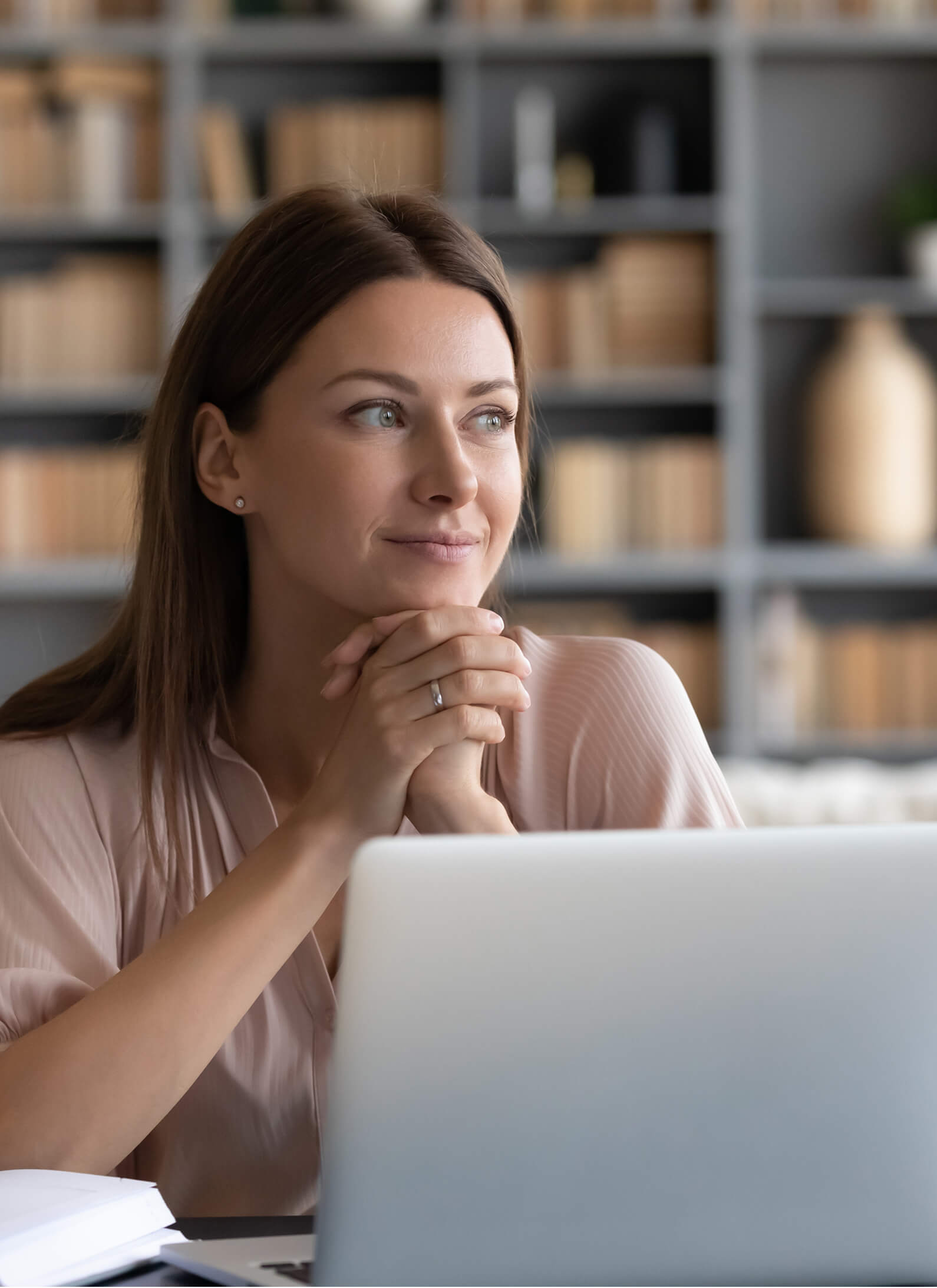 A woman sits at a desk with a laptop, hands clasped under her chin, looking thoughtfully to the side; shelves with books and decor are in the background.
