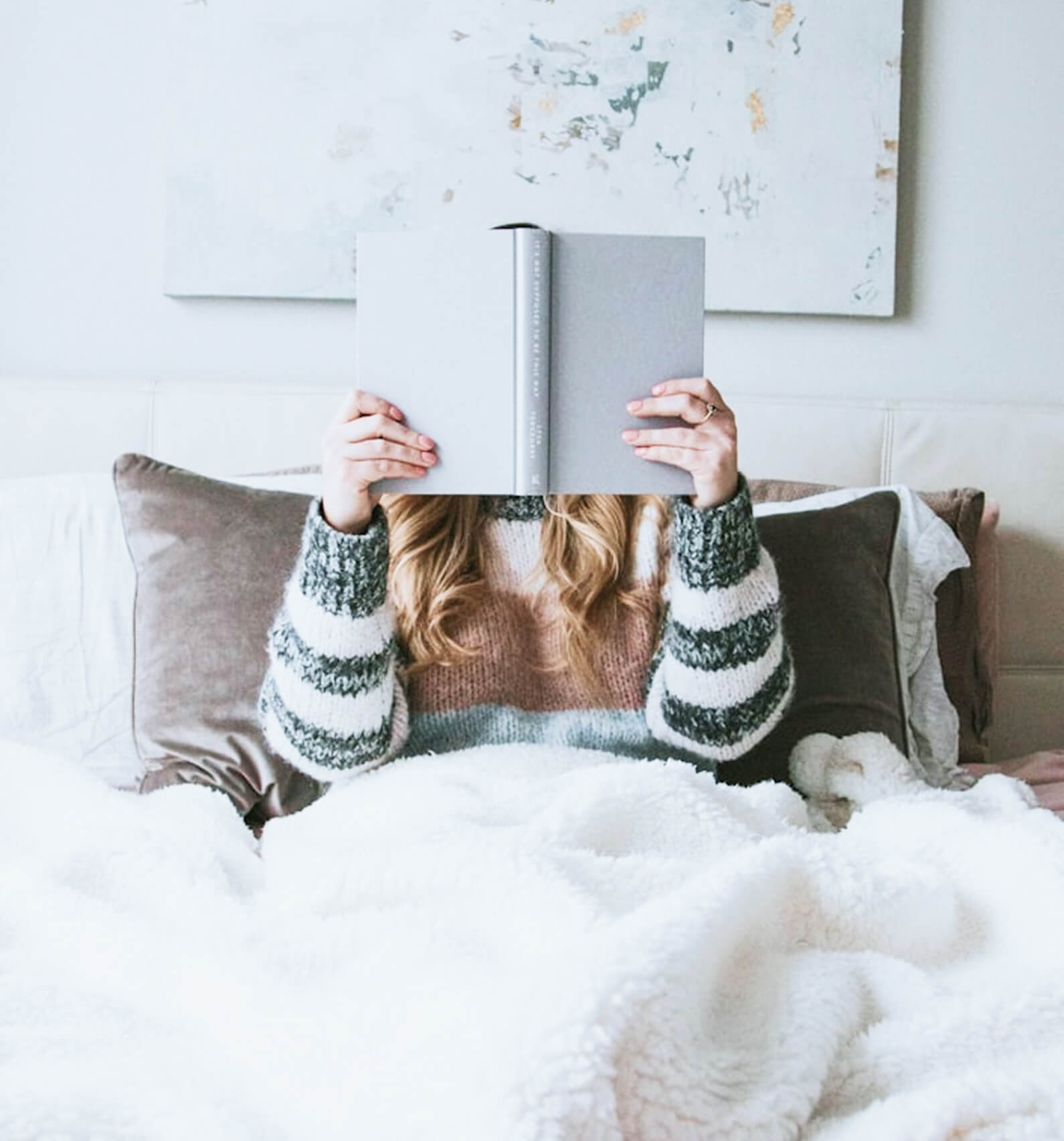 A person sits in bed under a white blanket, holding and reading an open book that covers their face.