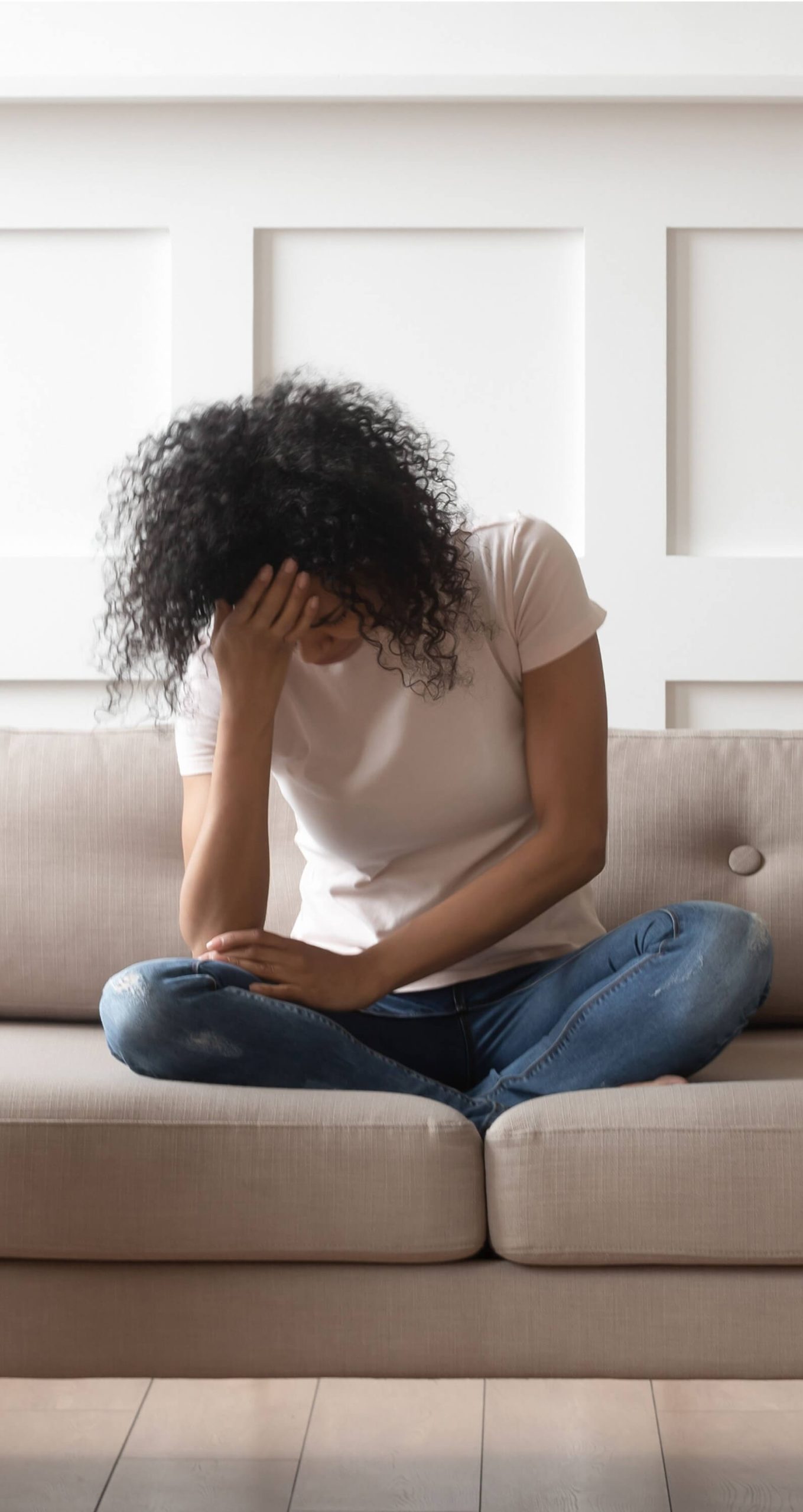 A person with curly hair sits cross-legged on a sofa, head down and hand covering their face, appearing distressed or upset.