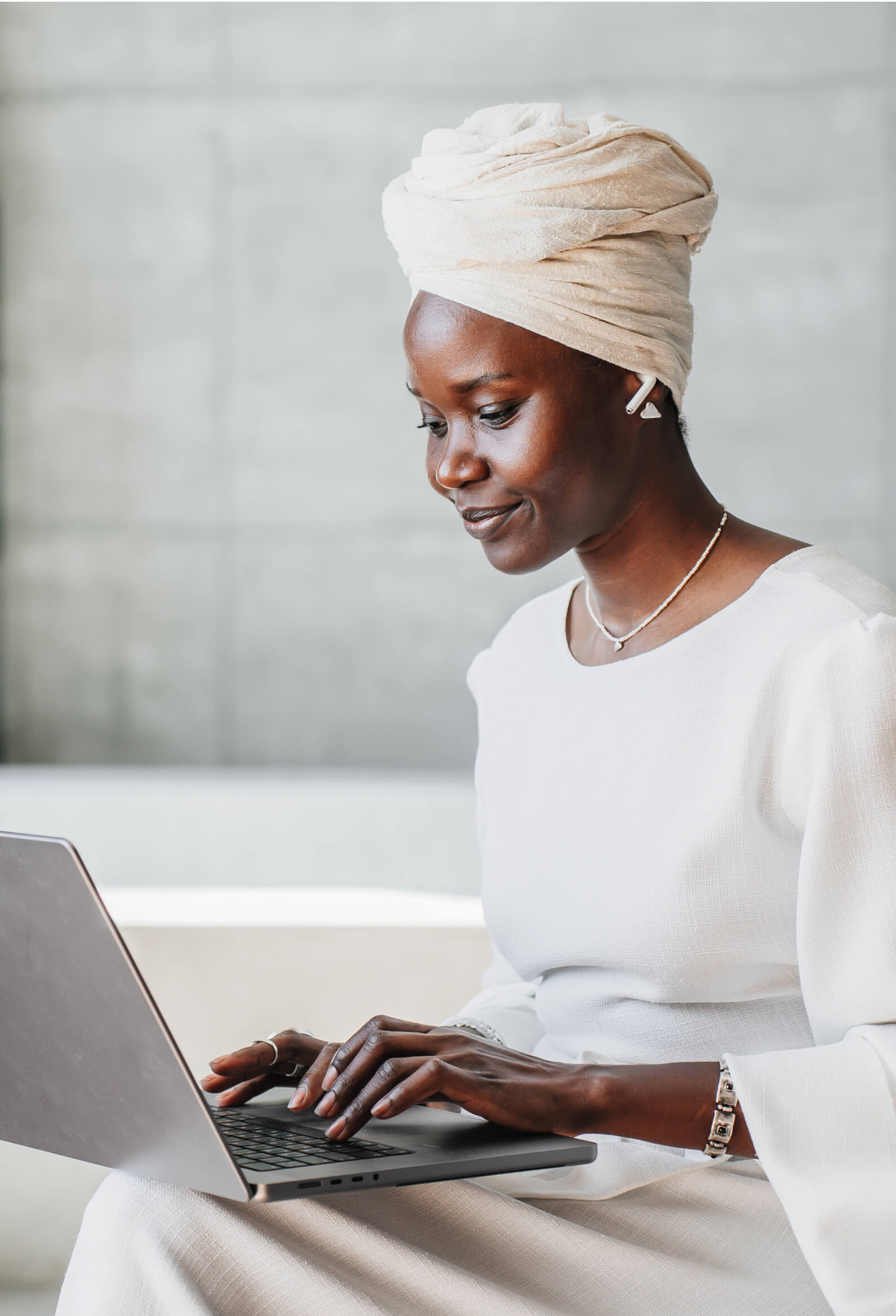 A woman in a white outfit and headwrap types on a laptop while wearing wireless earbuds, sitting indoors against a neutral background.