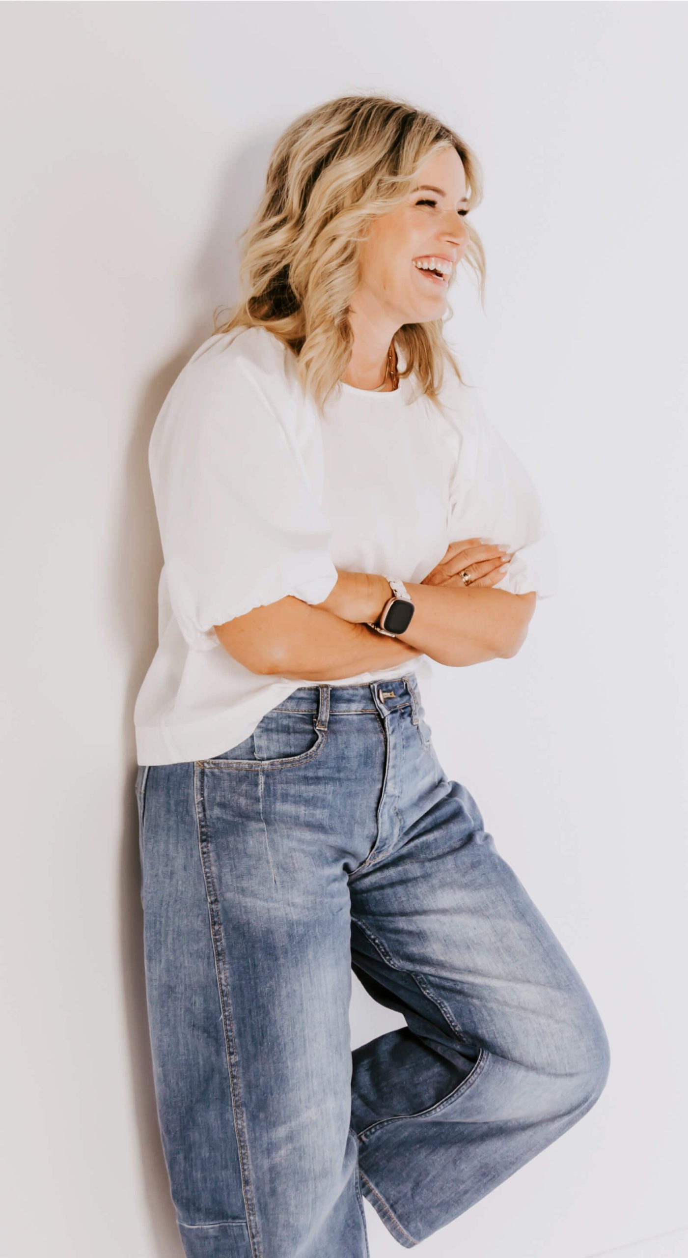 Person with shoulder-length blonde hair wearing a white shirt, blue jeans, and a smartwatch stands against a white wall with arms crossed, smiling and looking to the side.