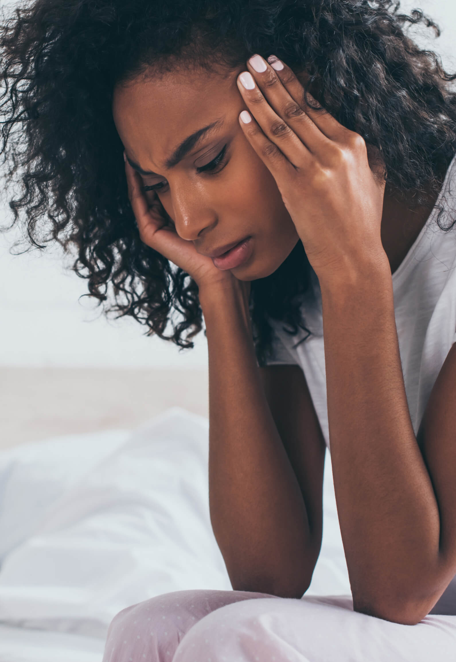 Woman sitting on a bed holding her head with both hands, appearing stressed or experiencing discomfort.