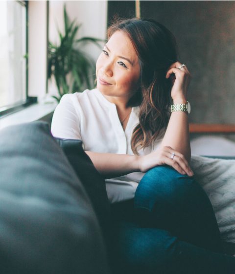 A woman sits on a couch near a window, resting her arm on the back of the couch and looking outside with a relaxed expression.