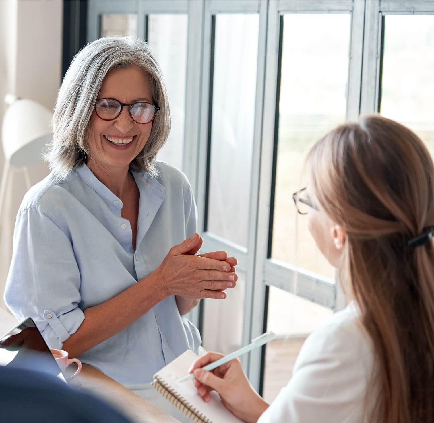 Two women sit by a window; one is smiling and gesturing while the other listens and takes notes in a notebook.