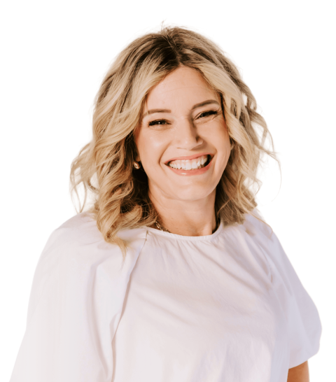 Woman with shoulder-length wavy blonde hair smiling, wearing a loose white top, standing against a plain background.