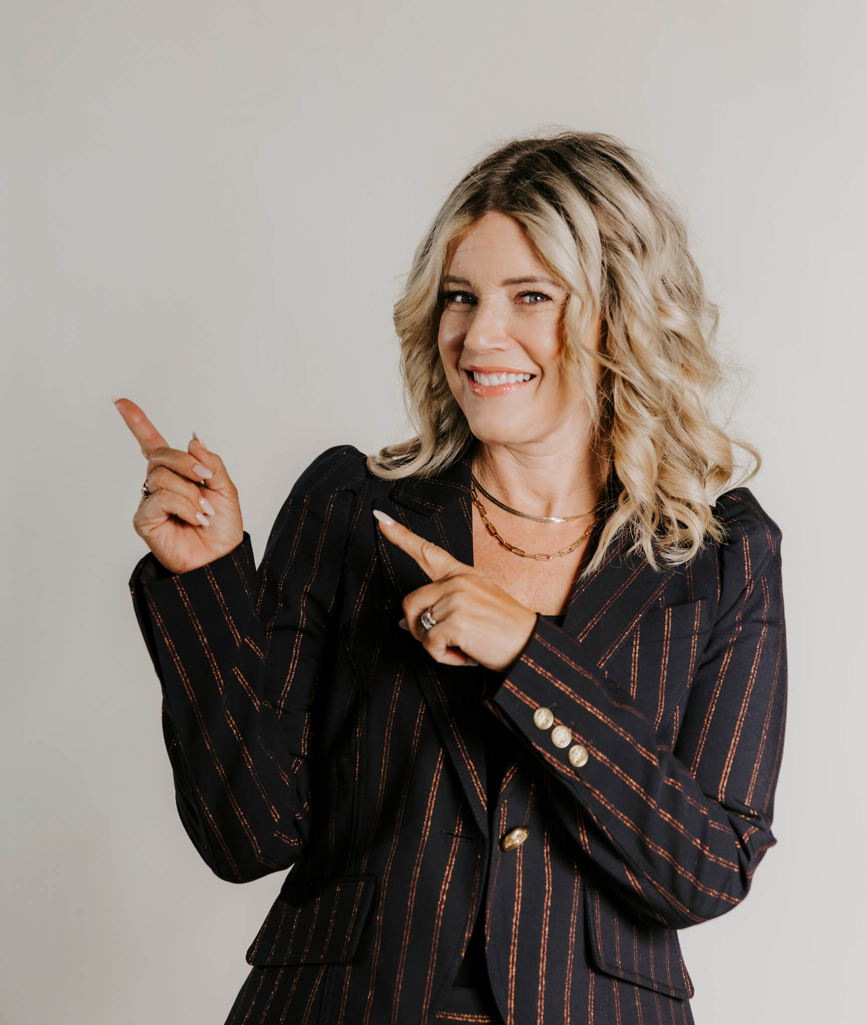 A woman with wavy blonde hair, wearing a pinstripe suit, smiles and points with both hands to her left against a light background.