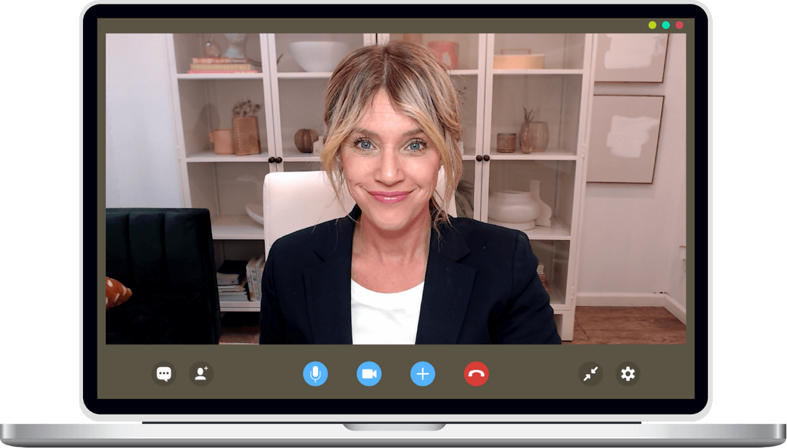 A woman wearing a blazer sits in a home office, smiling at the camera during a video call on a laptop screen.